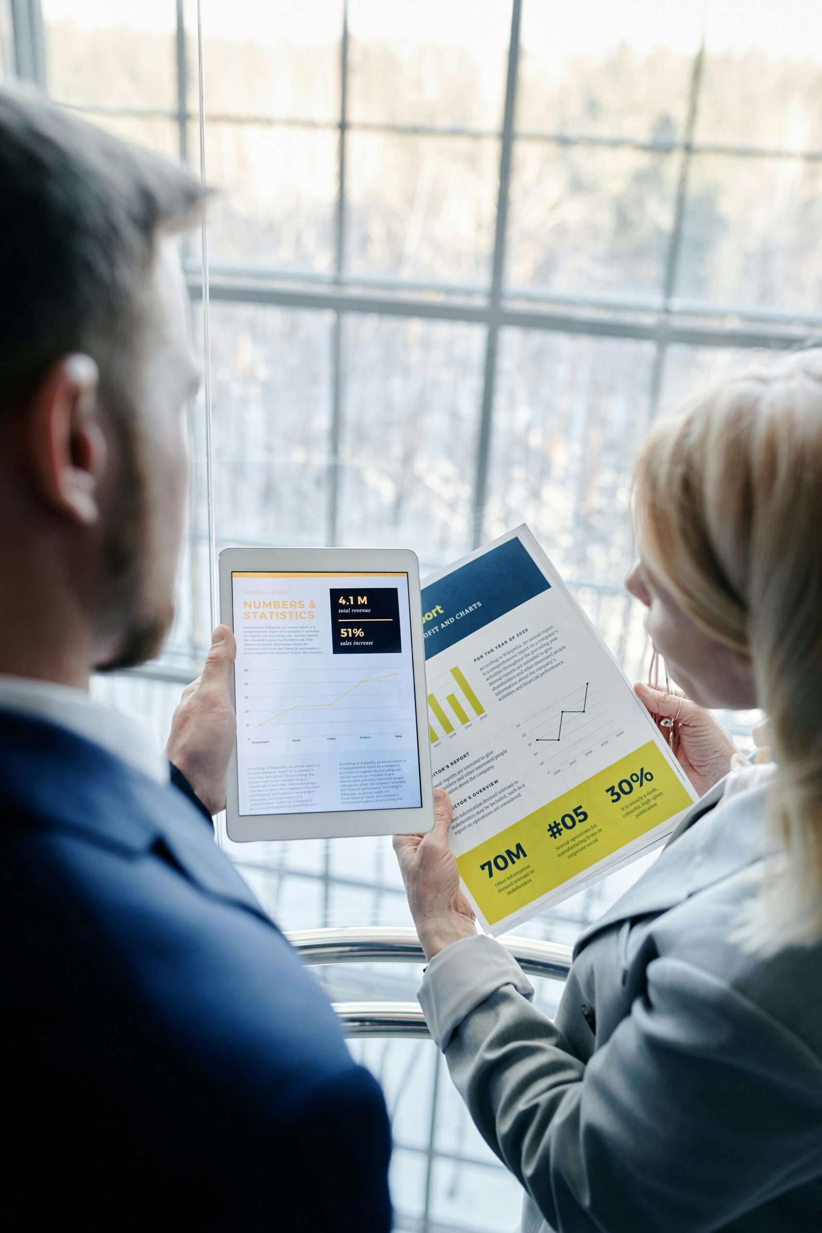 Two business professionals reviewing graphs and data on a tablet and paper report indoors.